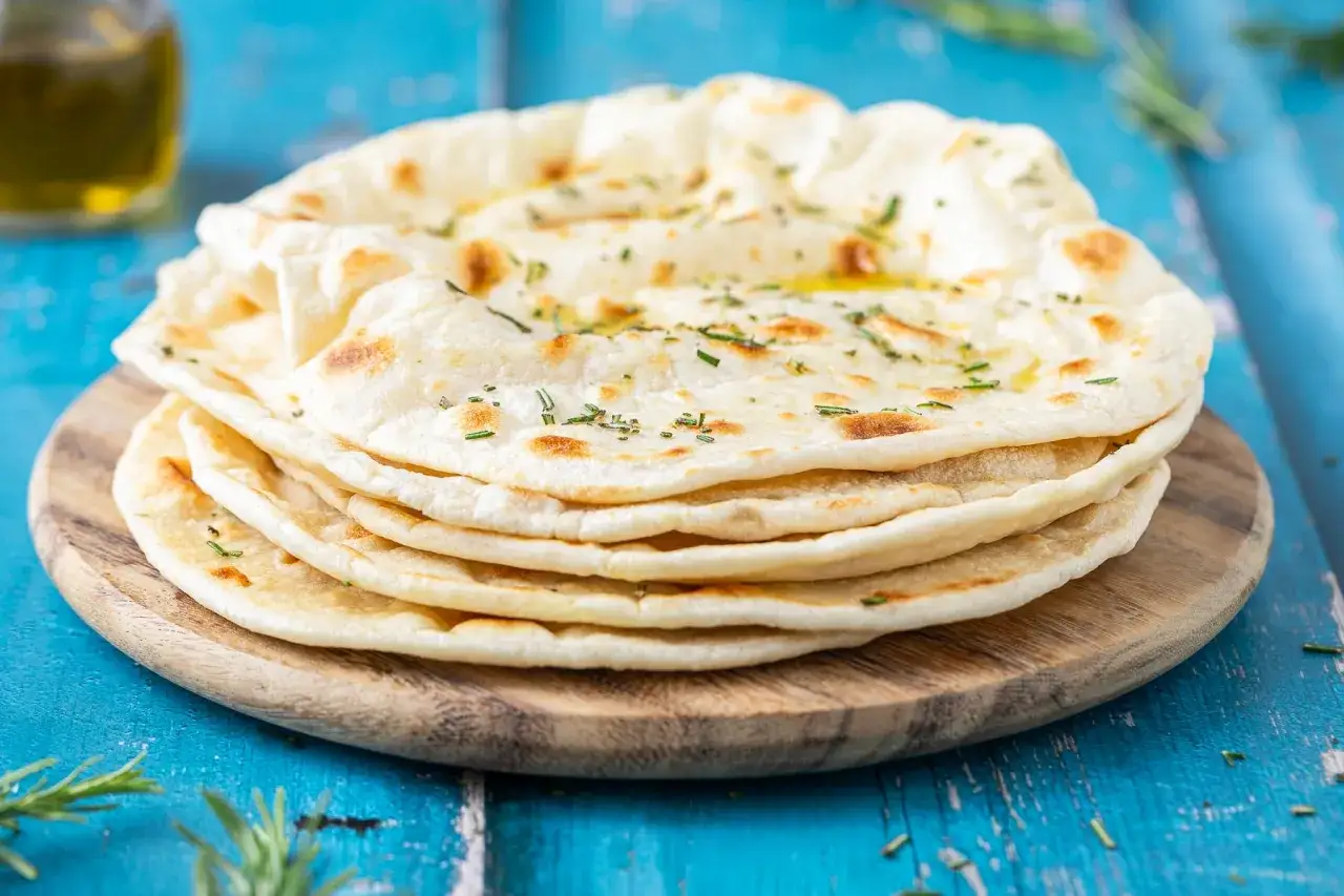 Pila di pane senza lievito, condito con erbe aromatiche e olio d'oliva, su un tagliere di legno.